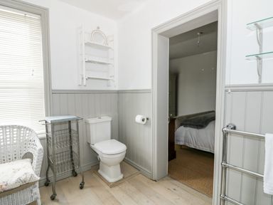 A bathroom with a toilet chair a metal cart and doorway to a bedroom at Troon Cottage in Hunstanton