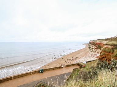 A beach with waves and rocks along the shoreline with grassy cliffs and a paved walkway at Troon Cottage in Hunstanton