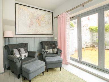 A sitting area with two armchairs two ottomans a floor lamp and a world map above wooden paneling beside glass doors with pink curtains at Troon Cottage in Hunstanton