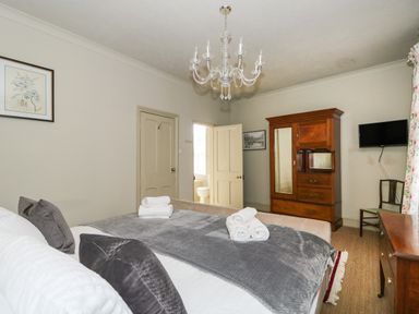 A bedroom with a bed with pillows and towels a wooden wardrobe a wall mounted television and a chandelier at Troon Cottage in Hunstanton