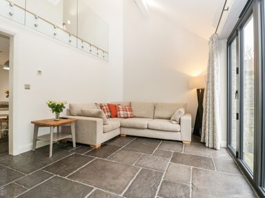 A living room with a beige sectional sofa a wooden side table with flowers floor lamp and sliding glass doors at Willow Barn in Shapwick