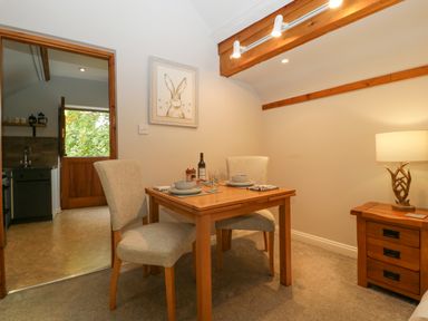 A dining area with a wooden table set for two and beige chairs next to a wooden nightstand with a lamp at The Coach House Apartment in Bredon