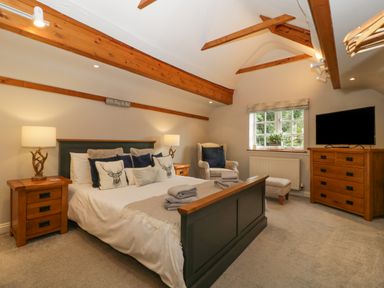 A bedroom with a double bed two bedside tables with lamps an armchair a window a chest of drawers with a TV and wooden ceiling beams at The Coach House Apartment in Bredon