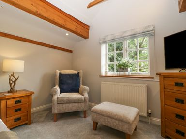 A small sitting area with an armchair a matching footstool a wooden side table with a lamp a window with floral decor and a wooden dresser with a TV at The Coach House Apartment in Bredon