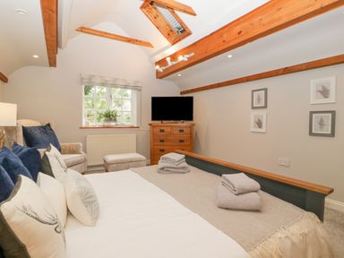 A bedroom with a bed, folded towels, a TV on a wooden dresser, armchair with ottoman, window, and framed pictures at The Coach House Apartment in Bredon