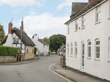 A street with white buildings including a thatched roof house and a pub sign with trees in the background at The Coach House Apartment in Bredon