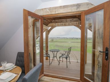 A dining area with table and chairs looking through open wooden doors to a covered balcony with metal table and chairs overlooking a grassy field at Hallgarth Farm Holiday Lodge in Peopleton
