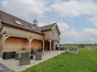 An outdoor patio with wicker chairs and table next to a wooden house with a grassy yard and children's play equipment at Hallgarth Farm Holiday Lodge in Peopleton