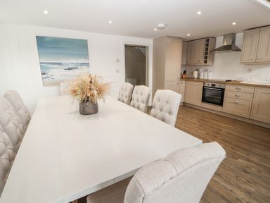 A dining area with a white table and tufted chairs next to a kitchen with beige cabinets and wooden floor at Sgubor in Valley