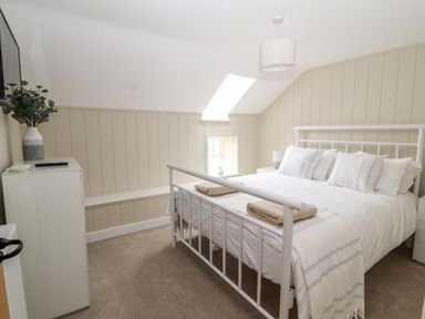 A bedroom with a white metal bed frame two folded towels on the bed a bedside table with a lamp a chest of drawers with a vase of leafy branches and a wall-mounted television at Sgubor in Valley