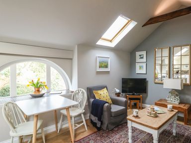 A living room with a small dining table and two chairs a gray armchair a coffee table with cups and pastries a television on a wooden stand and decorative mirrors at Little Brook in Nottington near Weymouth