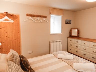 A bedroom with a bed with rolled towels and cushions a dresser with a mirror a window and wall hooks with hangers at Little Brook in Nottington near Weymouth