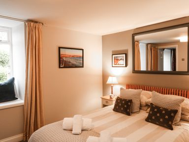 A bedroom with a double bed, pillows, folded towels, a side table with a lamp, framed pictures on walls, and a window with curtains at Little Brook in Nottington near Weymouth