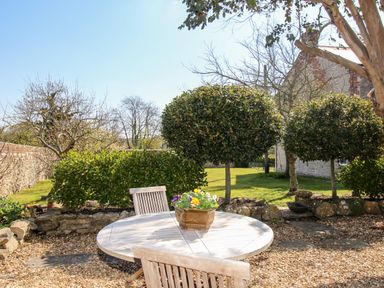 A garden with a round wooden table and chairs a flower pot on the table trimmed trees and a stone wall at Little Brook in Nottington near Weymouth