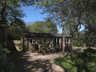 A wooden building surrounded by trees and gravel at Orchard Lodge in Bolney