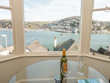 A glass table with a wine bottle and two wine glasses near windows overlooking a harbor with boats and houses at 4 Dartview in Dartmouth