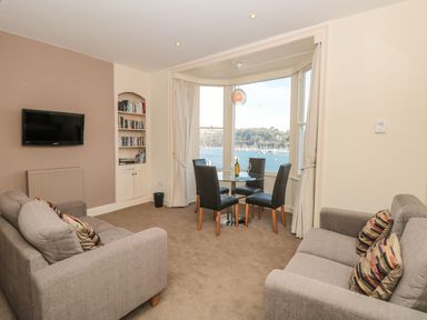 A living room with two grey sofas a wall-mounted television a built-in bookshelf and a dining table with four chairs by large windows overlooking a body of water at 4 Dartview in Dartmouth