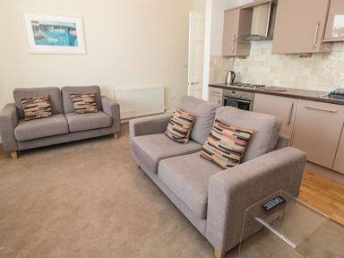 A living room with two gray sofas with patterned cushions a glass side table with a remote and a kitchen area with cabinets and stove at 4 Dartview in Dartmouth