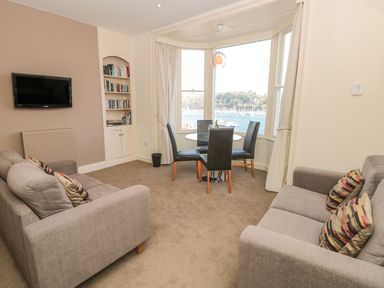 A living room with two gray sofas facing each other a wall-mounted TV a built-in bookshelf and a dining table with four chairs by a bay window overlooking water at 4 Dartview in Dartmouth