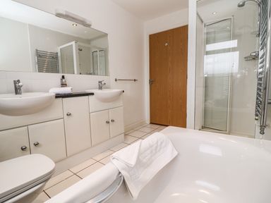 A bathroom with double sinks, a mirror, a bathtub with a towel, a shower with glass doors, and a wooden door at Redwood, Retallack in Retallack near St Columb Major