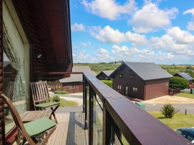 A balcony with two wooden chairs and a bench overlooking wooden houses and a rural landscape at Redwood Retallack in Retallack near St Columb Major