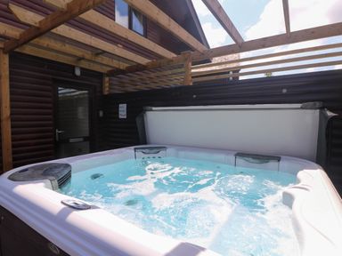 An outdoor hot tub with bubbling water under a wooden pergola next to a log cabin wall at Redwood Retallack in Retallack near St Columb Major
