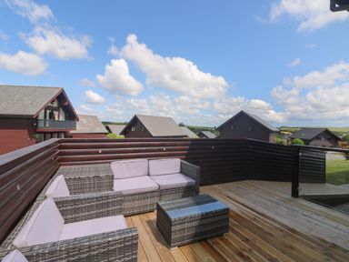 An outdoor wooden deck with wicker seating and a table surrounded by wooden fenced houses at Redwood Retallack Retallack near St Columb Major