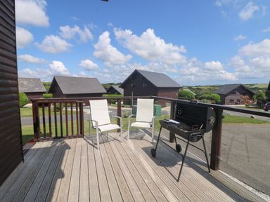 A wooden deck with two white chairs and a barbecue grill overlooking wooden cabins and green hills at Redwood Retallack in Retallack near St Columb Major