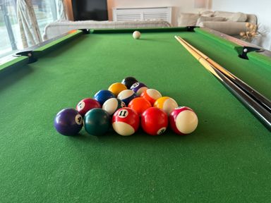 A pool table with balls racked in a triangle and two cue sticks resting on the side in a room with sofas