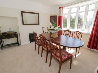 A dining room with a wooden table and six chairs near bay windows with red curtains at The Nook in Benllech