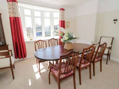 A dining room with a wooden table and six chairs near a bay window with red and cream curtains at The Nook in Benllech