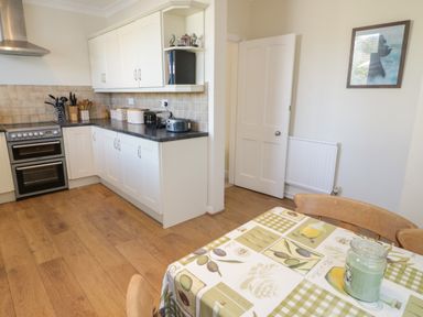 A kitchen with white cabinets a stove countertop appliances and a dining table with a tablecloth at The Nook in Benllech
