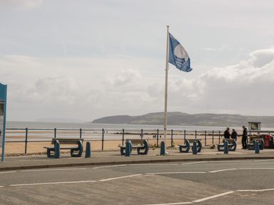 A beachfront promenade with benches a blue flag on a pole and people near the beach at The Nook in Benllech