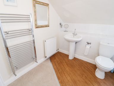 A bathroom with a toilet, pedestal sink, towel radiator, wall mirror, and wooden floor at The Nook in Benllech