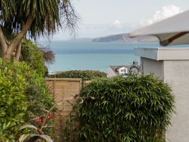 A garden with trees and bushes overlooking the sea with houses and hills in the distance at The Nook in Benllech