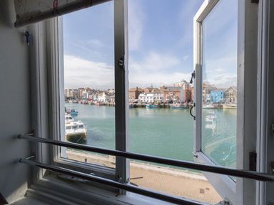 A view of boats docked in a harbor through an open window with buildings along the waterfront at Harbourside Cottage in Brewers Quay Harbour