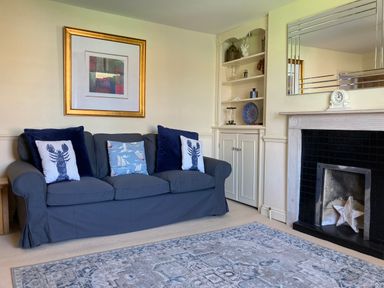 A sitting room with a grey sofa with blue and patterned cushions a framed abstract painting built in shelves and a fireplace at Harbourside Cottage in Brewers Quay Harbour