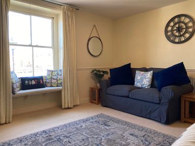 A living room with a grey sofa a round wall clock a round wall mirror and a window seat with cushions at Harbourside Cottage in Brewers Quay Harbour