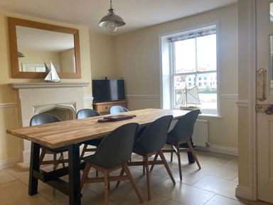 A dining room with a wooden table six chairs a fireplace mirror TV and a window with a model sailboat at Harbourside Cottage in Brewers Quay Harbour