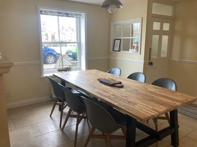 A dining room with a long wooden table and six gray chairs near a window with a model sailboat on the sill at Harbourside Cottage in Brewers Quay Harbour