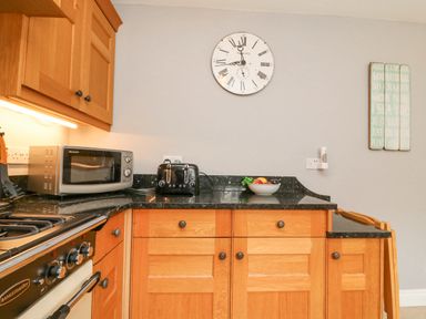 A kitchen counter with wooden cabinets a microwave toaster a bowl of fruit a clock on the wall and a gas stove at Harbourside Cottage in Brewers Quay Harbour
