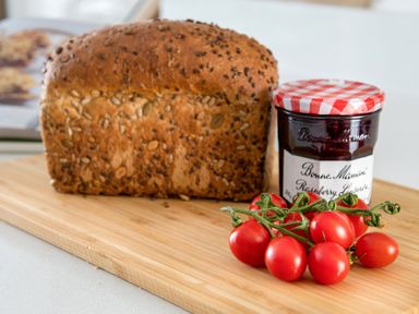 A loaf of seeded bread a jar of raspberry jam and a bunch of cherry tomatoes on a wooden cutting board