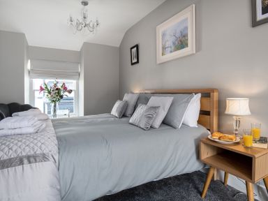 A bedroom with a double bed gray bedding pillows a wooden nightstand with a lamp plate of pastries and two glasses of orange juice a vase with flowers on the windowsill at The Old Sweet Shop in Conwy