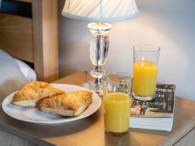 A bedside table with two glasses of orange juice a plate with pastries a book and a lamp at The Old Sweet Shop in Conwy