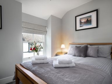 A bedroom with a wooden bed made with gray bedding and towels on top a window with blinds and flowers beside the bed at The Old Sweet Shop in Conwy