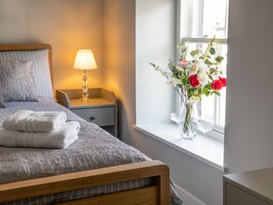 A bedroom with a wooden bed with folded white towels on a bedspread a bedside table with a lamp and a window sill with a vase of flowers at The Old Sweet Shop in Conwy