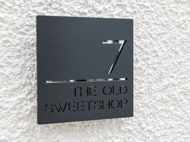 A black square house number sign with number 7 and text The Old Sweetshop on a white textured wall at The Old Sweet Shop in Conwy