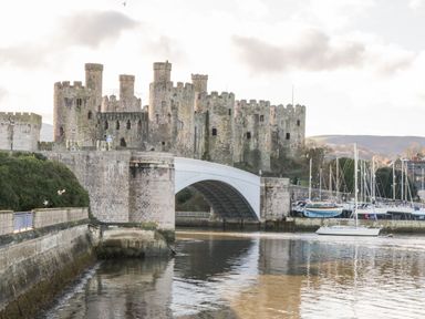 A stone castle with multiple towers beside a bridge over a body of water with boats docked near the castle at The Old Sweet Shop in Conwy