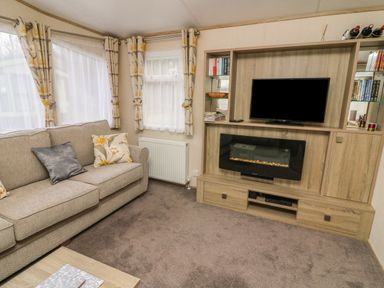 A living room with a sofa curtains and a TV on a wooden unit with an electric fireplace at 12A Borwick Lakes South Lakeland Leisure Village