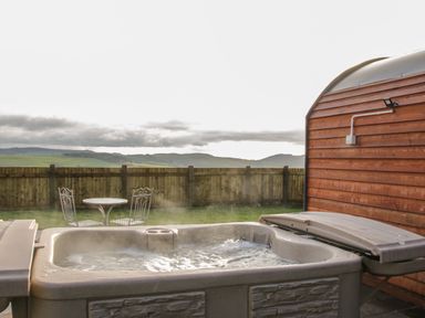An outdoor hot tub next to a wooden cabin with a metal table and two chairs set on grass at Stapeley Pod in White Grit near Chirbury